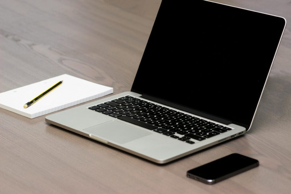 A clean and simple office desk setup featuring a laptop, smartphone, and notebook with pencil.