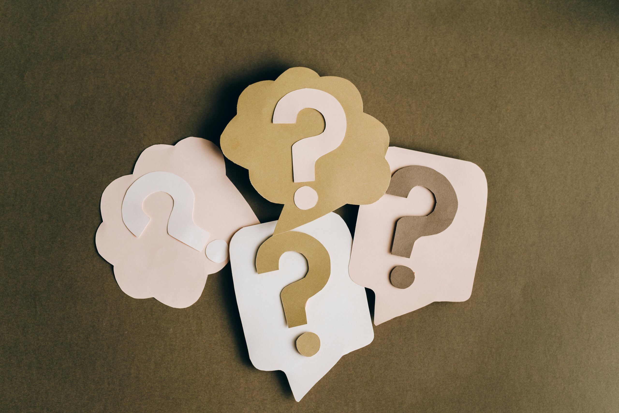 Four paper cutouts of question marks in beige and white on a brown surface, symbolizing inquiry and curiosity.